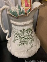 Front view of the white ceramic chamber pot showing scalloped rim and green floral decoration with a handle. No chips or cracks visible.