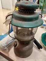 Vintage green Coleman lantern with glass globe, showing rust and dirt, photographed on a table.