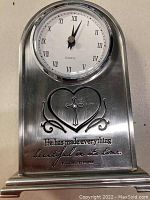 Front view of silver-colored metal mantle clock showing clock face with Roman numerals and religious heart and cross motif with inscription.