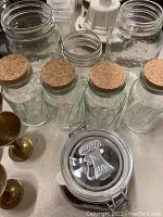 Photo showing vintage glass jars of various sizes with cork lids, two large mason jars without lids, brass goblets, and a glass jar with clamp lid