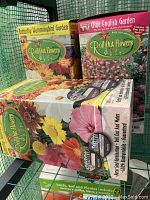 Three boxes of Roll Out Flowers garden seed mats on green wire rack, showing Buttercup Aluminumbird Garden, Olde English Garden, and a floral variety with sunflowers and pink blooms.