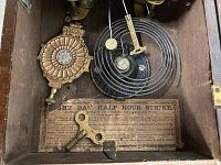 View inside the clock cabinet showing mechanical parts including a decorative brass pendulum bob, coiled spring, winding key, and an instruction label.
