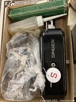 Photo shows the Singer buttonhole attachment unit along with a plastic oil bottle, some extra metal parts wrapped in plastic, and a green box for accessories.