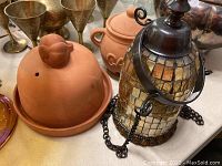 Photo showing terracotta stoneware covered butter or garlic dish with bird-shaped knob and a glass mosaic hanging lantern with metal accents and chain on a table with other glass and metal items.