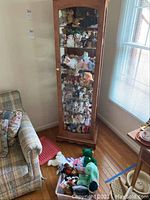 Full view of glass wooden cabinet holding multiple shelves of Beanie Babies, plus additional Beanie Babies in a pink plastic container on the floor next to the cabinet.