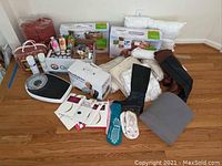 Photo showing the full lot arranged on hardwood floor including blankets, scale, bottles of lotions and shower gels, sock pairs, pantyhose, Tempur-Pedic boxed pillow, neck pillows, women's boots and a boxed massager.
