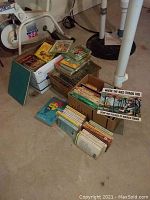 Photo showing three boxes of various children's and young adult books on a basement floor, including bright covers and stacks of books.
