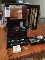 Open wooden jewelry box showing mirrored door, hanging necklaces on side panel, cubby with watches and bracelets, and lower drawer containing trays of earrings and brooches
