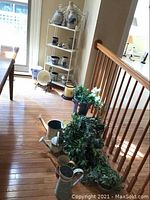 Photo showing metal watering cans, faux plants and a candle on wooden floor near staircase