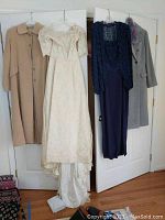 Full view of two gowns and two coats hanging on a wall: a white wedding gown, a navy blue lace gown, a beige coat, and a gray coat.