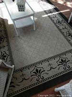 View of full rectangular outdoor rug with beige background, black floral decorative border and diamond pattern central field, placed under patio furniture.