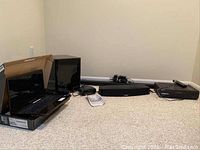 Wide photo showing all items in the lot placed on a carpeted floor including laptop, speakers, clock radio, and VHS player.