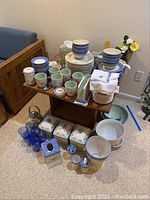 Overall view of the Pfaltzgraff dishware set including plates, bowls, mugs, kitchen accessories arranged on and under a wooden table.