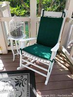 Photo showing white wooden rocking chair with green cushions and white wood table with bird cage on top on a porch.
