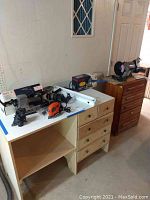 Overview of various power tools on white shelves and wooden drawer unit, showing sanders, boxed drill and joiner, bench grinder, and fluorescent light fixture.