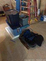 Stack of five plastic storage bins in dark blue and gray, with lids, also black and blue garment bags in front on floor.