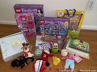 Wide shot showing all items for sale including LEGO Friends sets, Barbie doll, assorted plush toys, books, wrapping paper, and photo albums arranged on the floor.