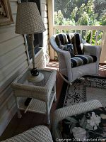 White wicker side table next to rocker chair, with lamp on top, porch setting.