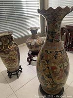 View showing three porcelain floor vases of varied sizes and shapes with painted art details, all placed near a window with blinds. Two vases on wooden stands.