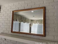 Large rectangular wall mirror mounted against a white brick wall, reflecting window treatments and ceiling lights.
