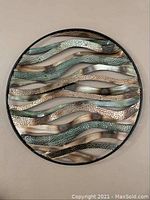 Front view showing circular metal wall art with wavy horizontal strips in silver, bronze, and green patina tones with varied textures.