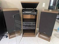 Full view of central wooden cabinet with record player on top and karaoke player plus amplifier below, flanked by two large Techics speakers with black fabric covers.