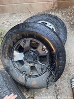 Four 265/70R16 tires on original 16-inch Toyota Tacoma wheels coated in black Plasti Dip, showing weathering and mud, stacked outdoors on ground.