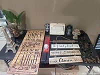 Overview of multiple decorative wall art pieces and items on a table including wooden plaques with quotes, metal crosses, framed art, Eiffel Tower sculpture, and metal bird hanging.