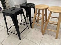 Four stools arranged in a row on tiled floor: 2 black metal stools and 2 wooden stools, clearly showing different materials and styles.