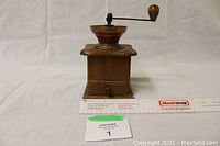 Full front view of a small vintage wooden spice grinder with a brass bowl and a metal crank handle with wooden knob on top, placed on a white cloth background.