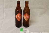 Two identical amber-colored vintage glass soda bottles with diamond-shaped orange and white labels reading 'Orange Crush Bottle'. Bottles are vertically ridged and empty.