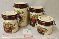 Four off-white round ceramic jars with metal clasps, each painted with a fruit basket design, displayed together with a ruler for scale.