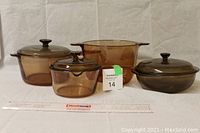 Six pieces of amber colored Pyrex glass bakeware including two pots with lids, one saucepan with lid, and two bowls with handles, arranged with a ruler for scale