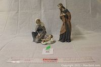 Set of three ceramic figurines showing Jesus lying on a cloth, Mary kneeling, and Joseph standing with cloak, placed on neutral grey backdrop with measuring ruler for scale.