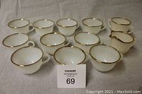 Photo showing 10 white tea cups and 1 creamer, 1 sugar bowl with gold rims arranged on grey fabric.