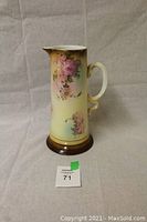 Full view of vintage off-white ceramic pitcher with a floral pattern and brown base on a neutral background with seller lot number.