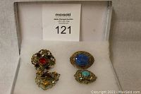 Four vintage brooches arranged on white fabric inside a box, showing front of brooches with various colored stones and intricate filigree metal work.