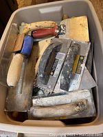 Wide view of a bin containing multiple drywall tools including trowels, putty knives and spreaders with different handle types and sizes, showing wear.