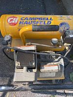 Full top and side views showing yellow cylindrical tanks with Campbell Hausfeld logo, grey motor housing, attached hoses and gauges, mounted on black frame with handle.