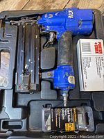 Blue pneumatic brad nailer with black grip and metal magazine, showing wear inside carrying case with box of Porter-Cable brad nails and package of staples.
