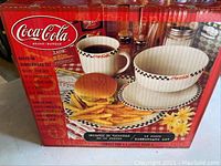 Front view of the boxed Coca-Cola dinnerware set showing the dinner plate, bowl, and mug with food and beverage.