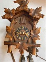 Front view of used wooden coo coo clock with carved leaves and bird detail on top, showing clock face with Roman numerals and hands.
