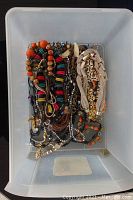 Top-down image showing multiple colorful beaded necklaces, fabric necklaces, metal chains and bracelets jumbled inside a plastic container.