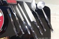 Nine Cutco utensils with black handles laid out: two forks, serrated spreader, three knives, two ladles
