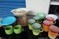 Photo showing various Fire King mugs in green and amber shades with diamond quilted design along with colorful small bowls and a plastic blue lid container.