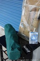 View of bundled green camping chair inside carrying bag, secured with black strap, shown alongside a metal frame in packaging and an accessory pack with cable lock.