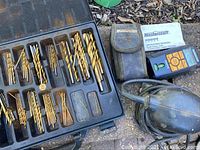 Box of assorted gold-colored metal drill bits organized in a black plastic case, a used black corded palm sander, and a blue and black Mastercraft laser level with its manual and soft carrying case.