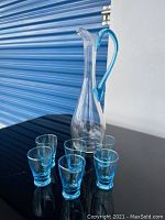 Photo of the full retro glassware set on a black surface: one tall clear glass pitcher with blue handle and six blue-tinted glasses arranged around it.