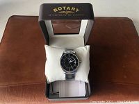 Rotary wristwatch with black dial displayed on a white cushion inside the branded box, showing face and bracelet details.