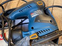 Side view of Ryobi sander showing blue and black body with orange accents and product label with specifications.